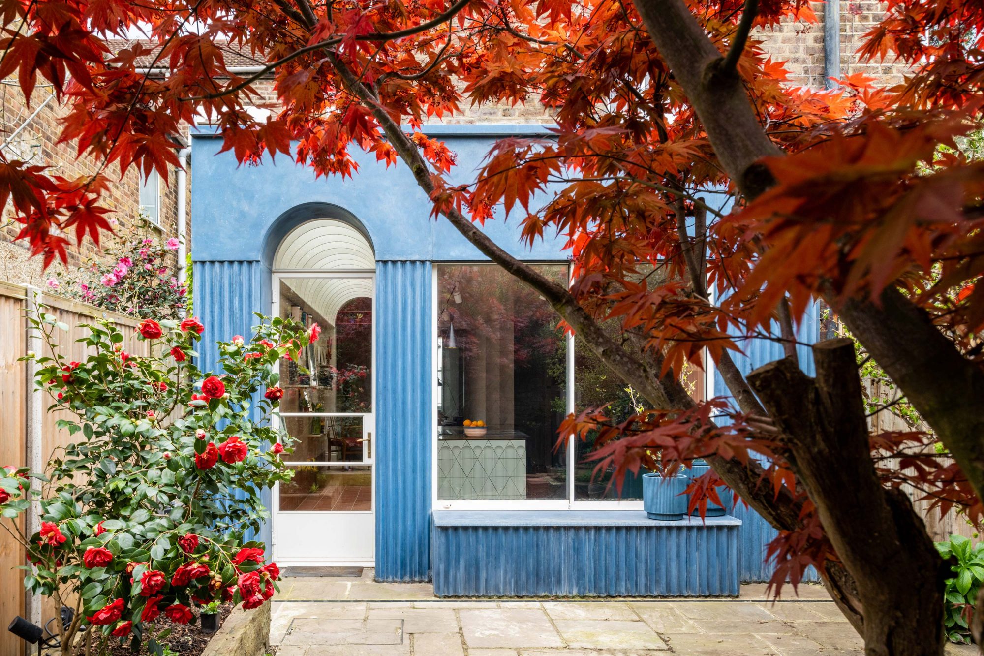 Exterior view of a London terrace by BVDS with bespoke blue corrugated concrete paneling, white arched glazing, a black Critall-style door, a built-in sitting bench, and a small red-leaved tree.