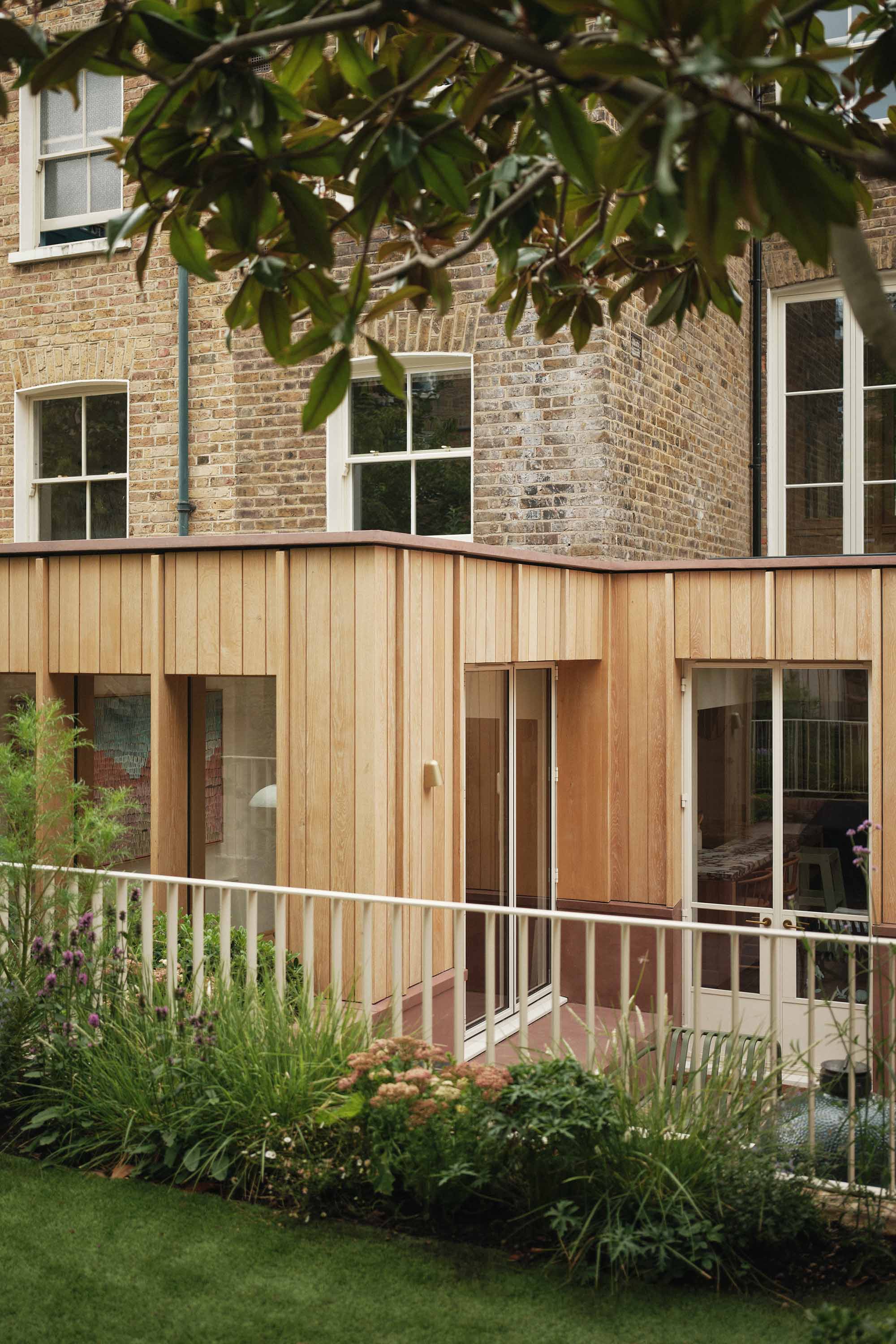 Lauriston raised garden view with metal railing balustrade, patio, outdoor kitchen, pink concrete detailing, oak panelling, rear extension, and planted landscaping.