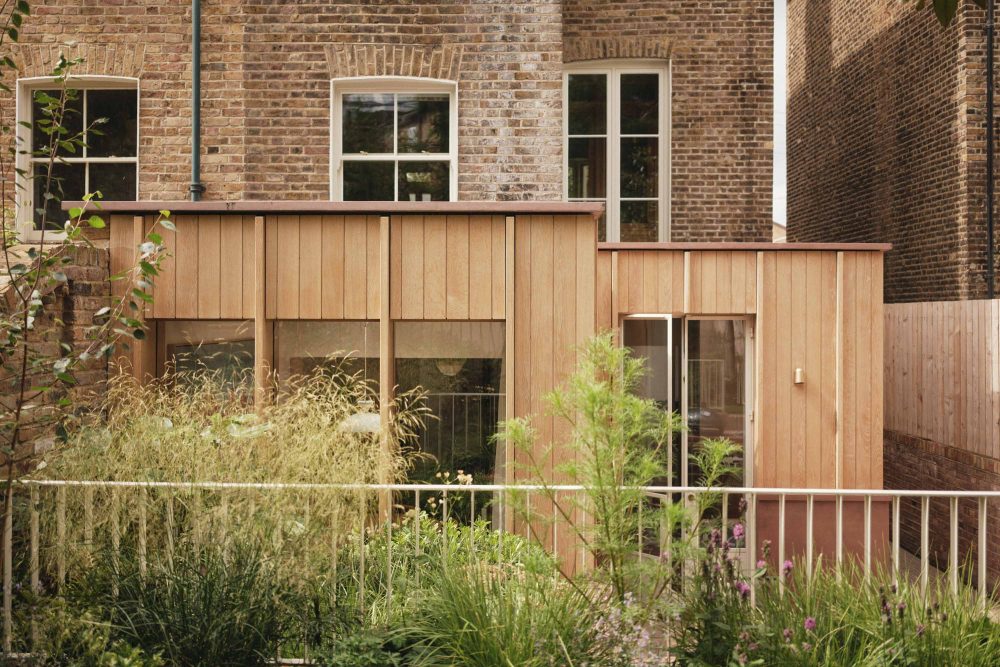 Lauriston raised garden view with metal railing balustrade, patio, outdoor kitchen, pink concrete detailing, oak panelling, rear extension, and planted areas.