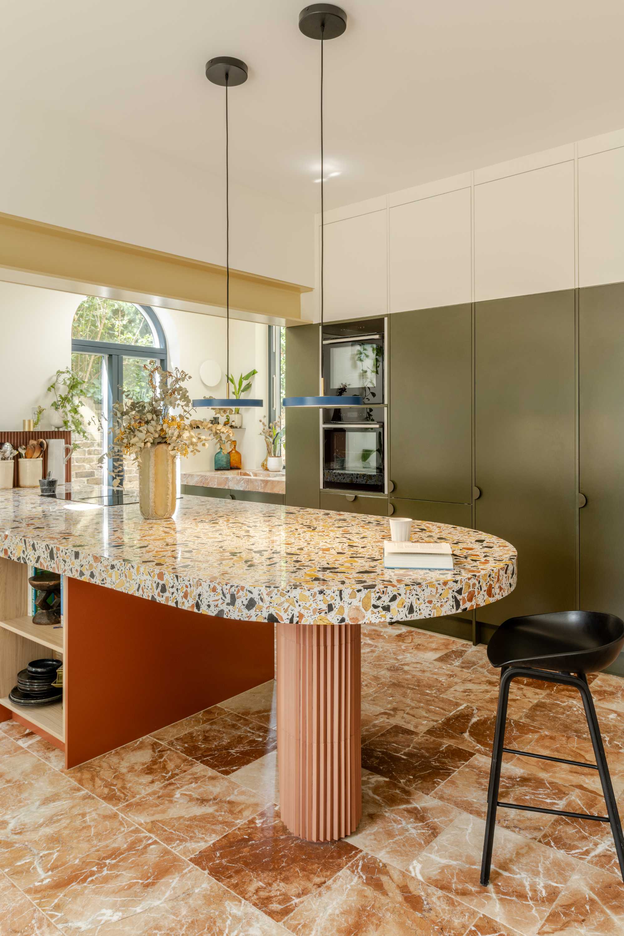 Modern kitchen extension with curved terrazzo island, olive-green cabinetry, and natural light from arched window.