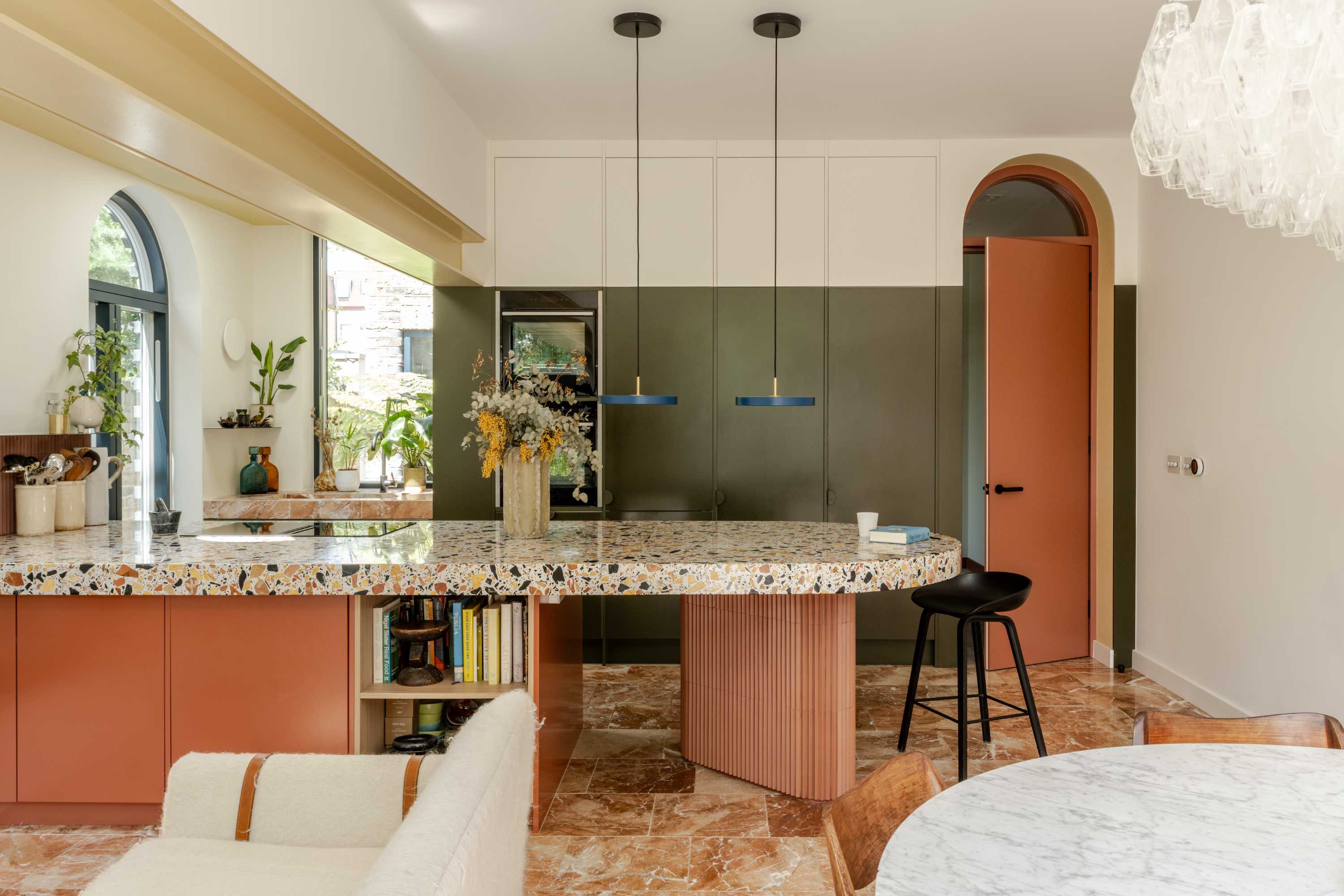 Open plan kitchen extension with terrazzo island, olive cabinetry, and coral accents. Natural light from arched window.