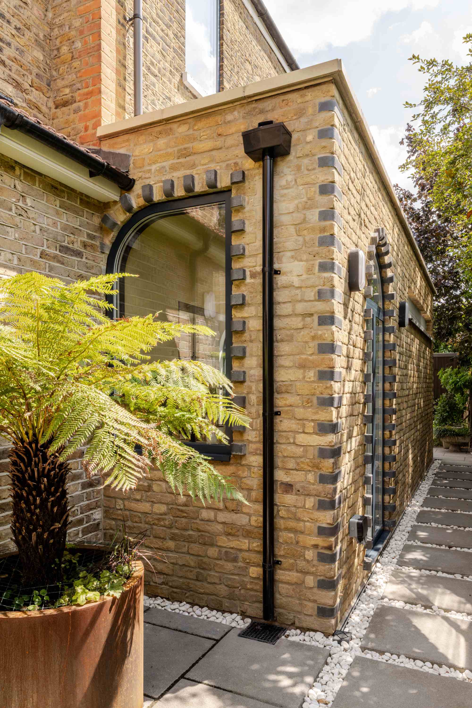 Contemporary stone garden room extension with arched black-framed window and decorative brick detailing, creating bright ope.